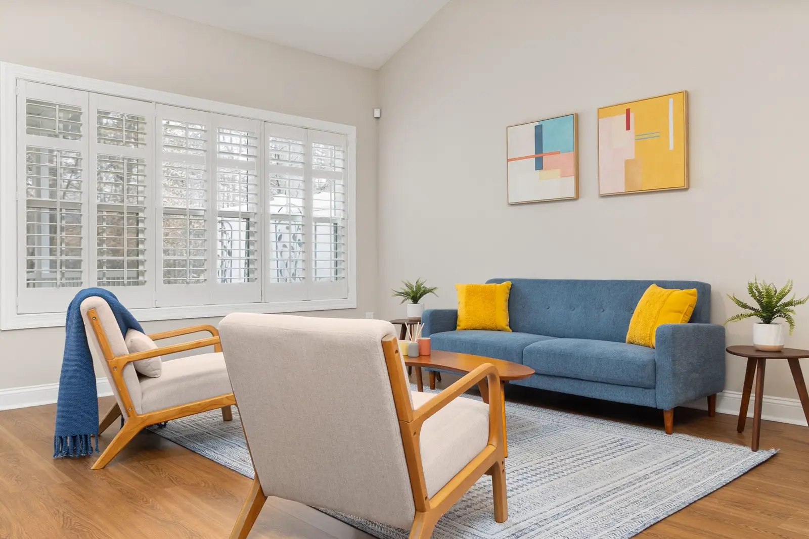 A bright living room with a blue sofa, wooden chairs and a sunlit shuttered window